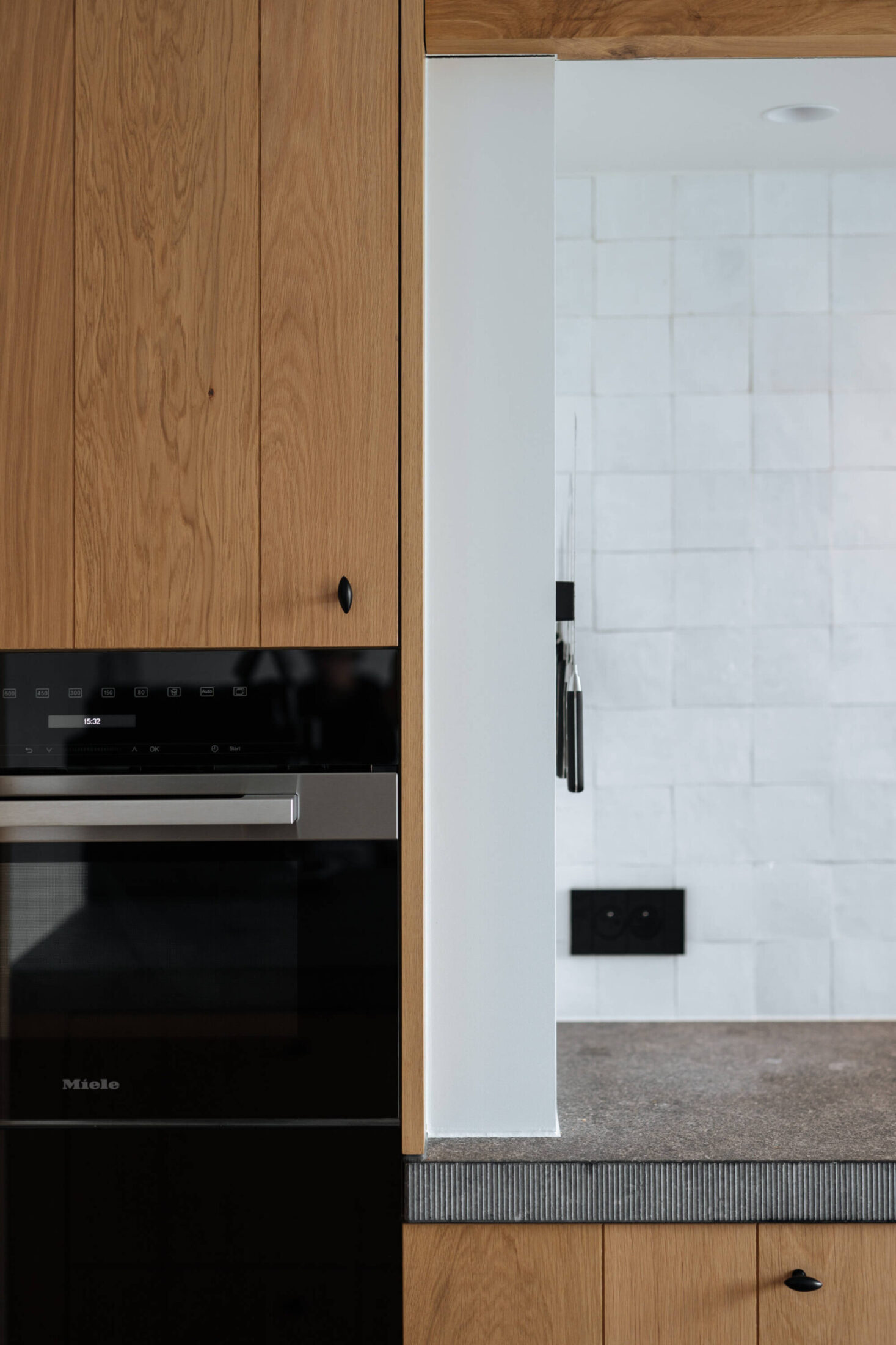 Detail van een landelijke keuken met eiken hoge kast, zwarte oven en witte wandtegels in Roeselare, Kaster Keukens