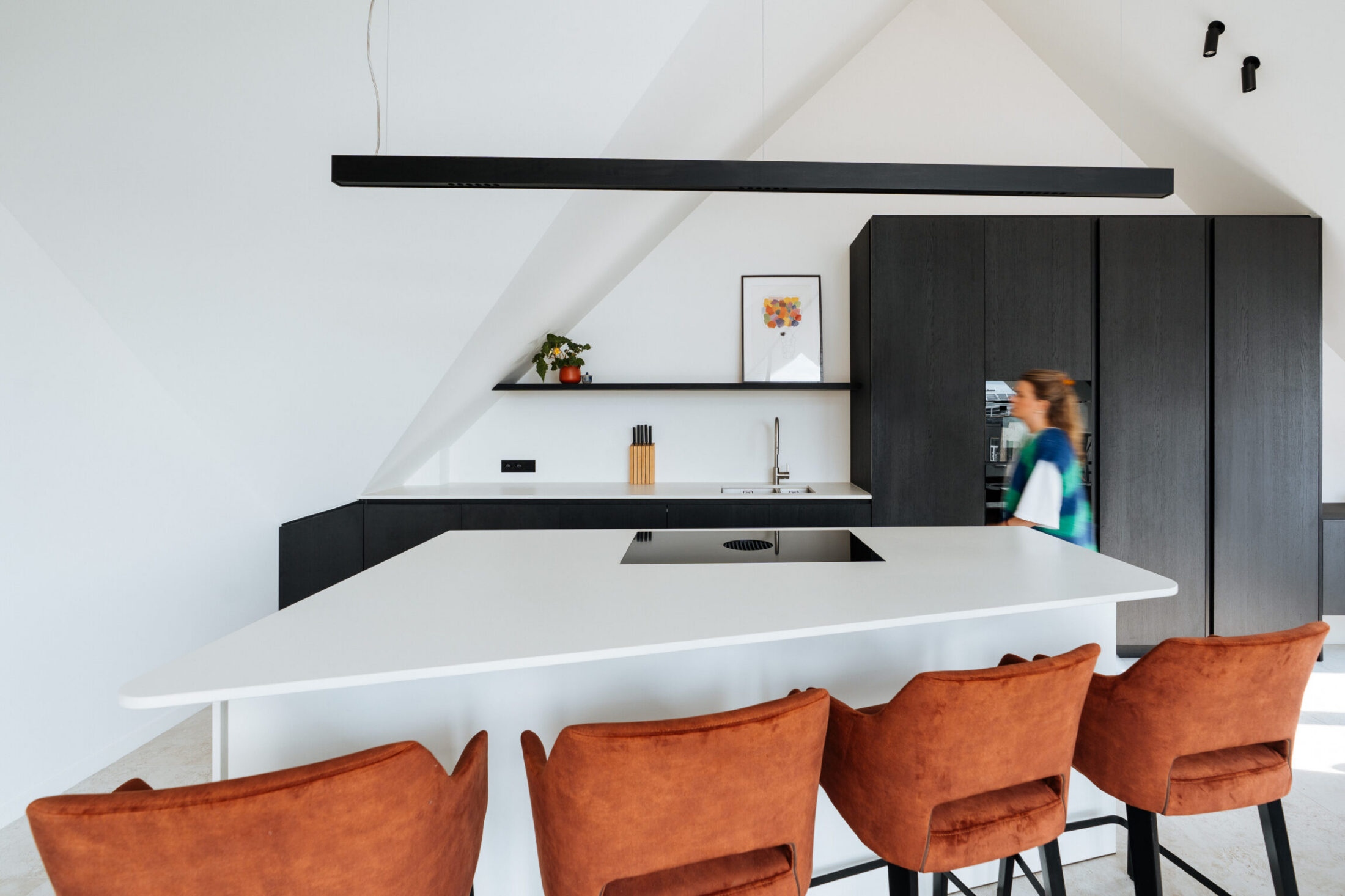 Moderne keuken met zwarte houten fronten, wit eiland en oranje barkrukken onder schuin dak in Knokke-Heist, Kaster Keukens