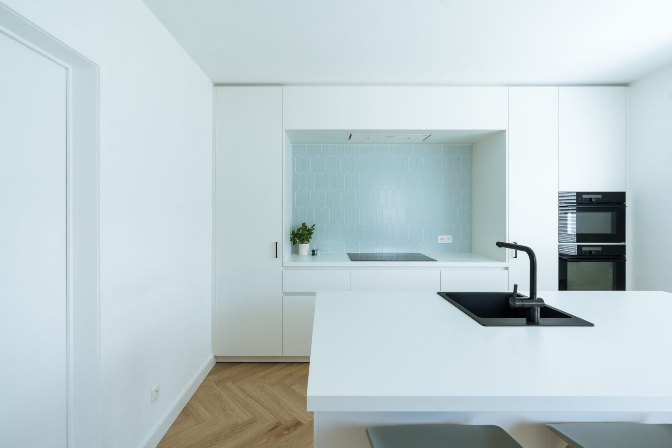 Moderne keuken met witte fronten, kookeiland met zwarte spoelbak en lichtblauwe wandtegels in Moorslede, Kaster Keukens
