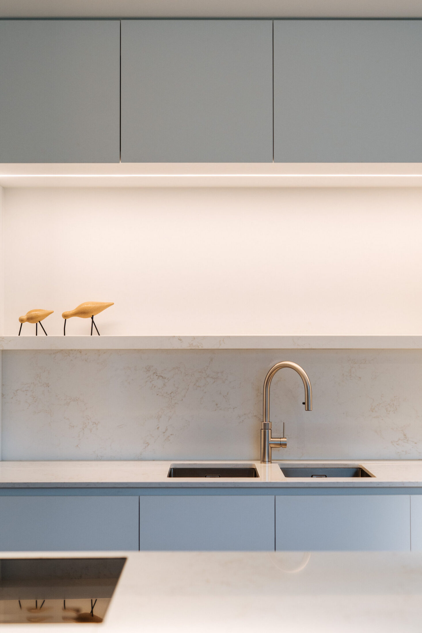 Detail van een moderne keuken met grijze bovenkasten, verlichte nis en koperkleurige kraan in Drongen, Kaster Keukens