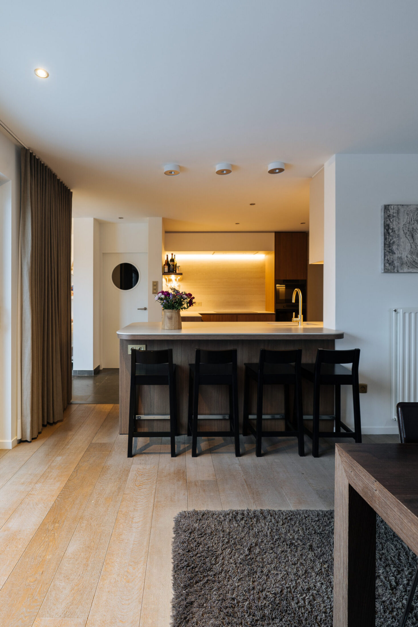 Moderne keuken met houten fronten, kookeiland en zwarte barkrukken, gezien vanuit de woonkamer in Roeselare, Kaster Keukens