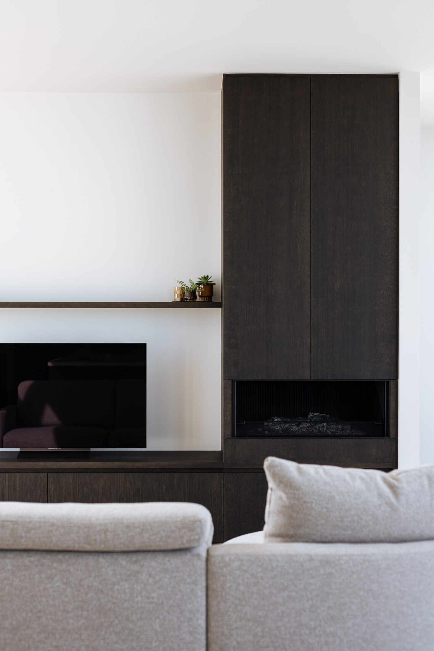 Tv-meubel op maat in donker hout met hoge kolom en haard achter een lichte zetel in Lendelede, Kaster Keukens