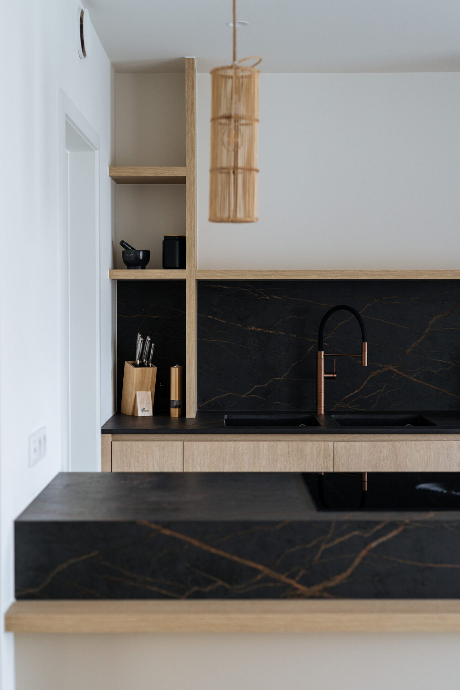 Moderne keuken met lichte houten fronten, zwart werkblad met gouden aders en rieten hanglamp in Knokke-Heist, Kaster Keukens