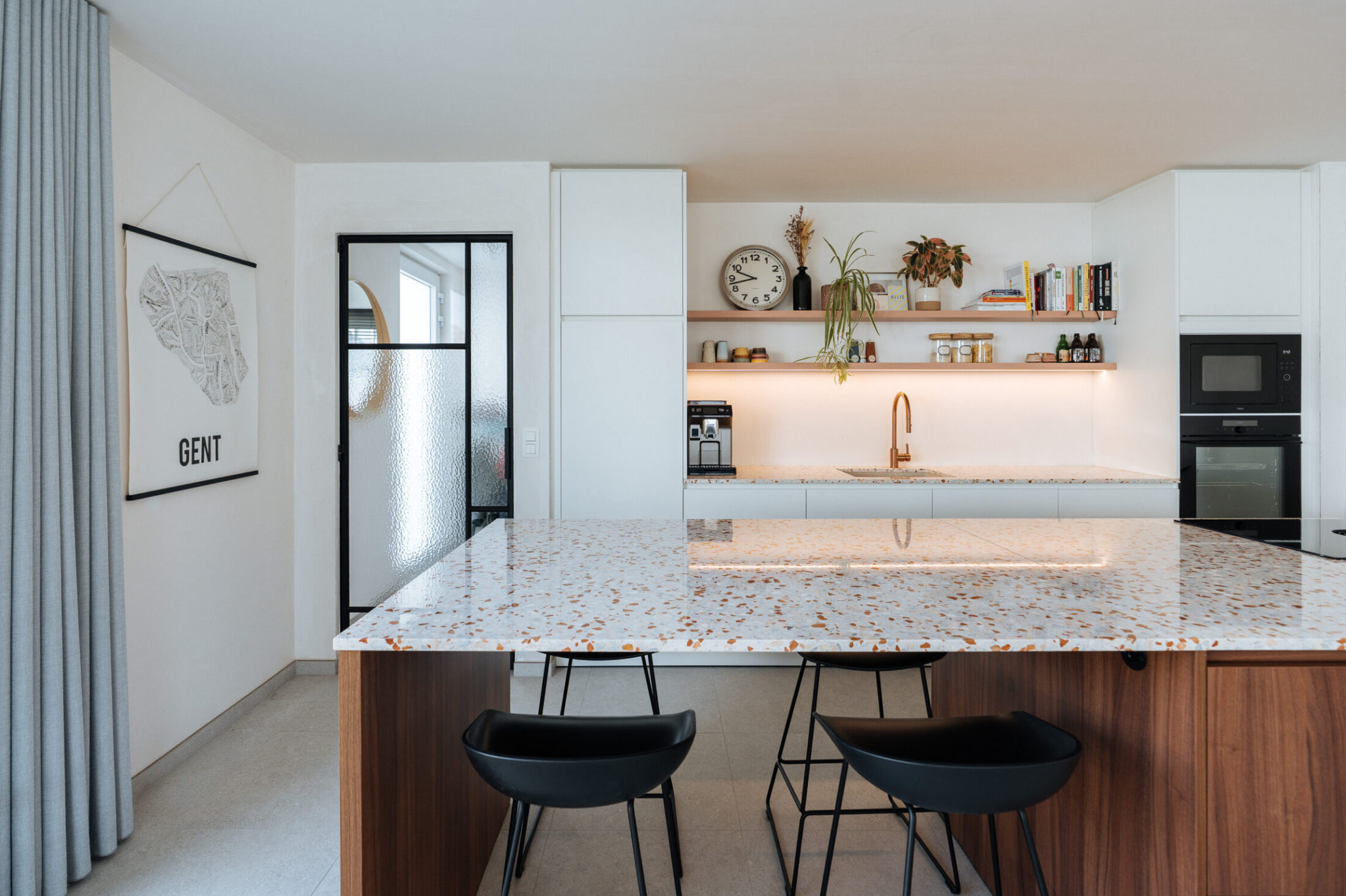 Moderne keuken met terrazzo eiland, notelaar onderkast, zwarte barkrukken en witte kastenwand in Merelbeke, Kaster Keukens
