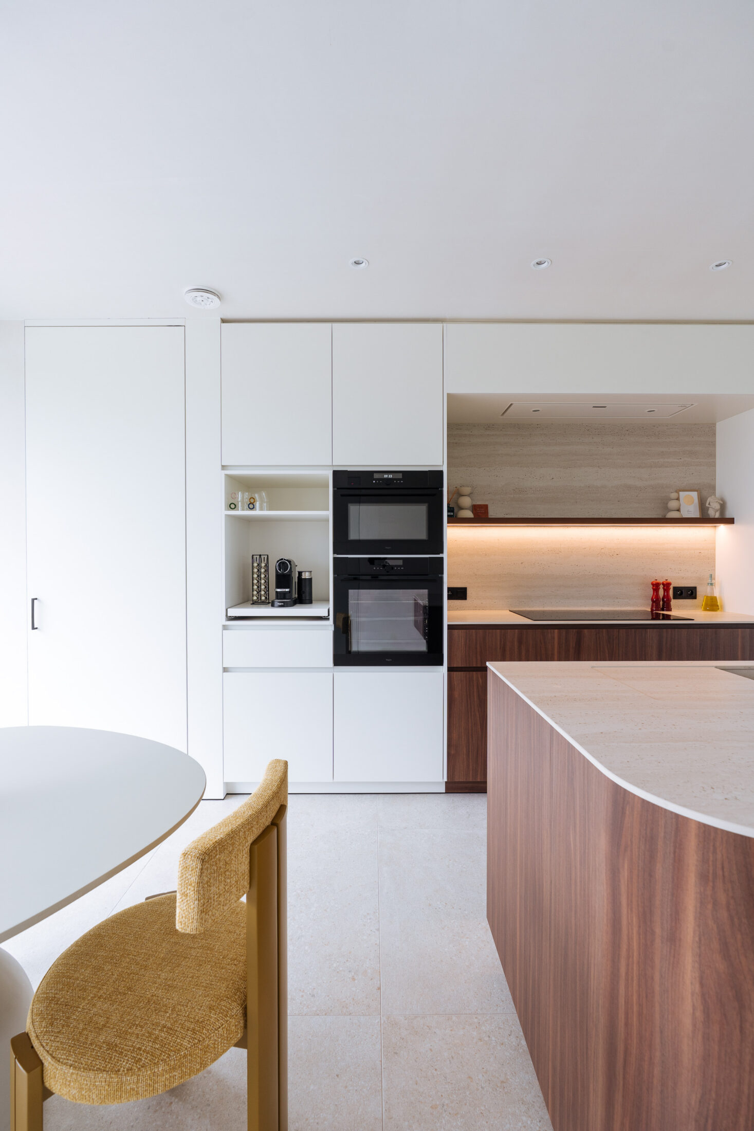 Moderne keuken met witte hoge kasten, ingebouwde ovens en gele stoel aan de eettafel in Hooglede, Kaster Keukens