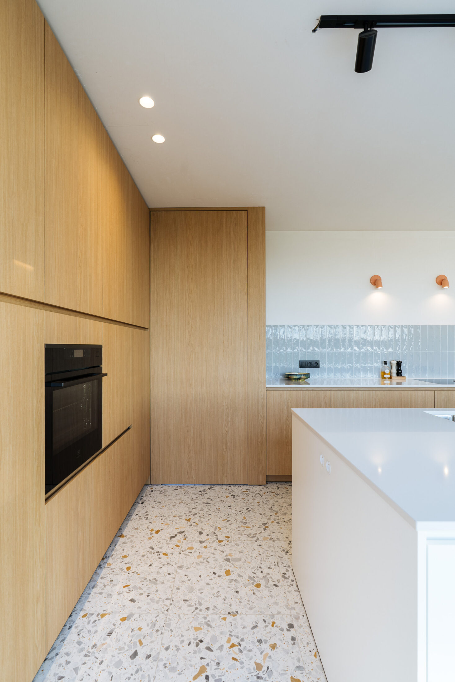 Moderne keuken met hoge eiken kastenwand, inbouwoven en wit eiland op terrazzovloer in Torhout, Kaster Keukens