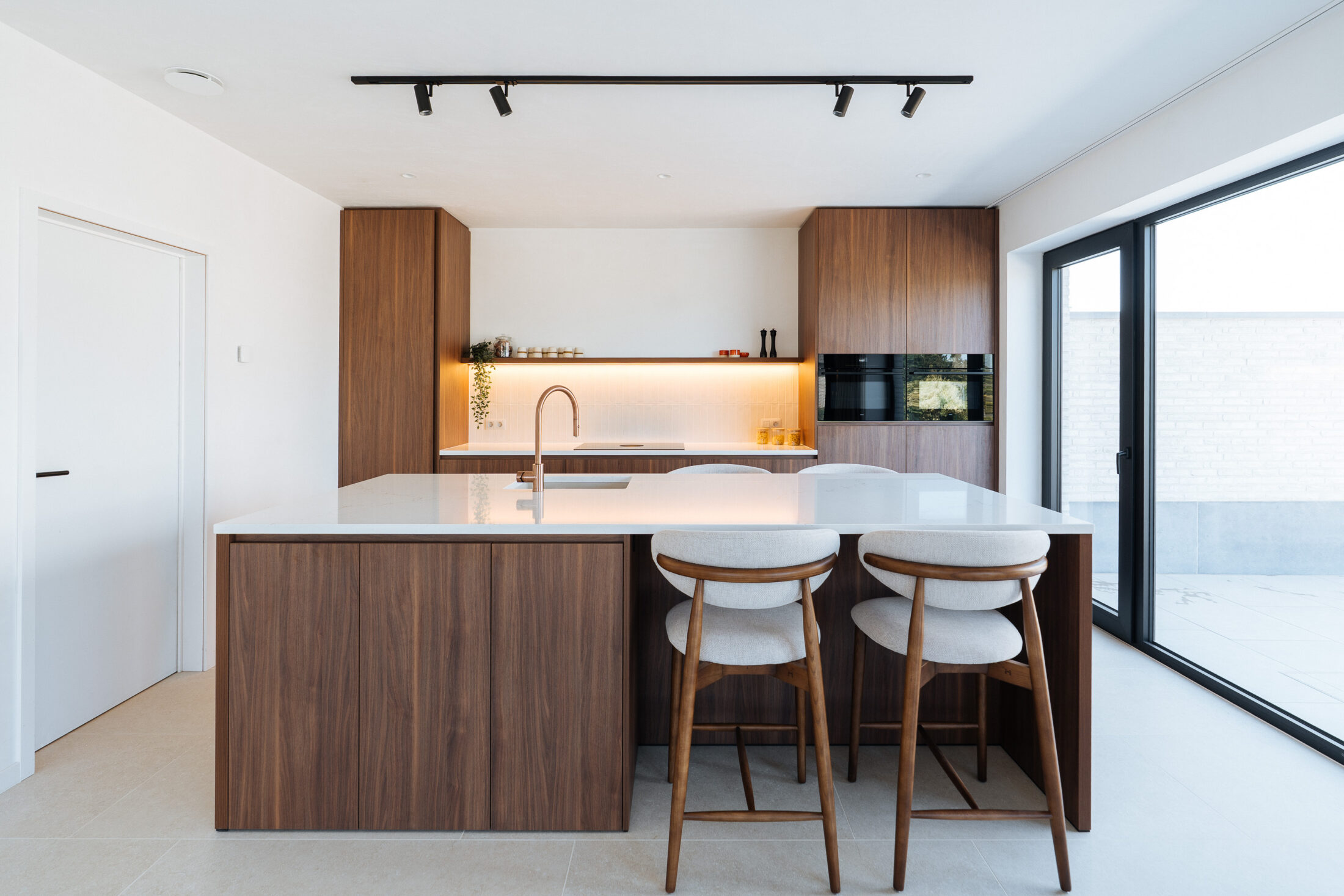 Moderne keuken met notelaarlook fronten, wit composiet werkblad, koperkleurige kraan en lichte barkrukken met houten onderstel, Kaster Keukens.