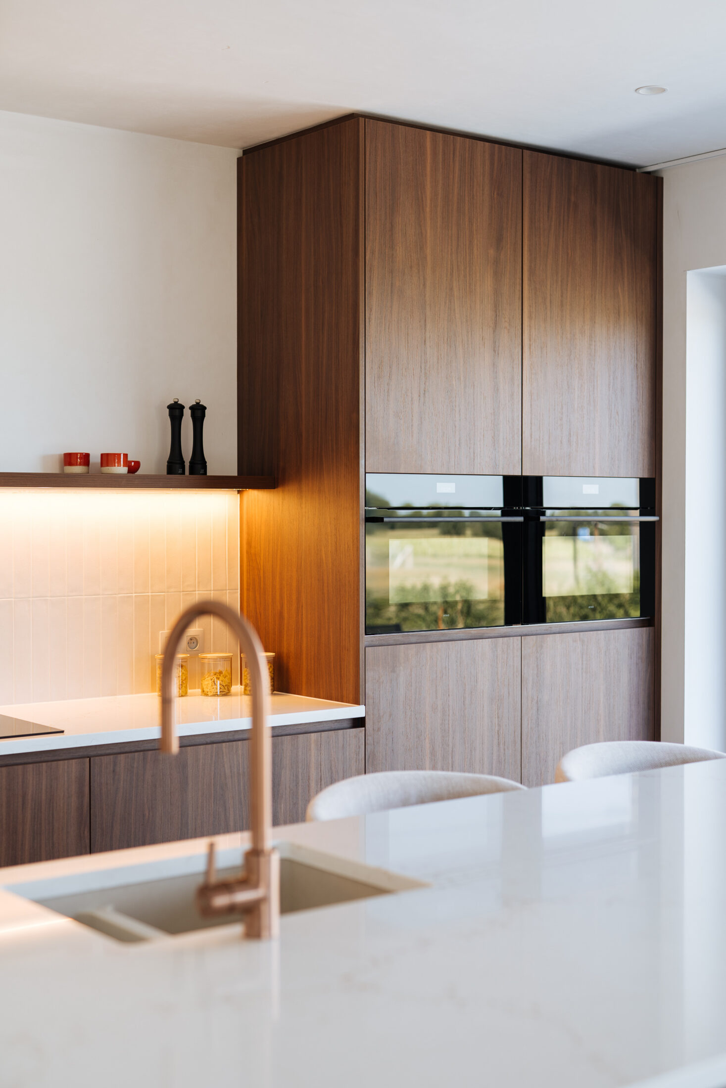 Moderne keuken met notelaarlook fronten, wit composiet werkblad, koperkleurige kraan en lichte barkrukken met houten onderstel, Kaster Keukens.