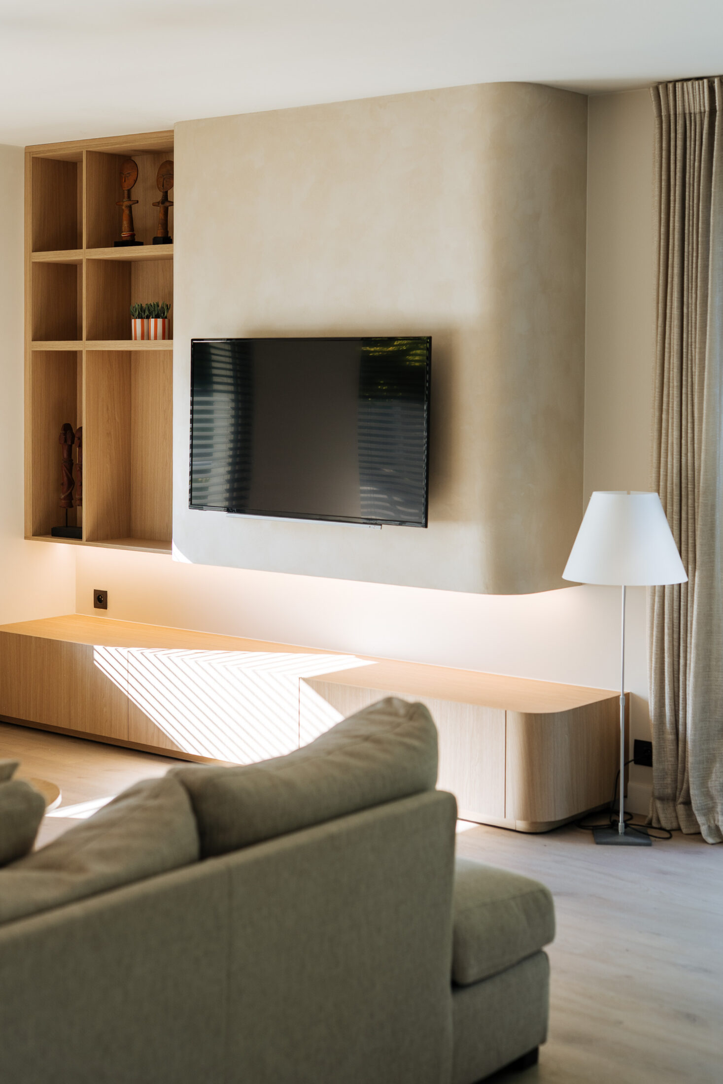 Tv-meubel op maat in lichte eikenlook met afgeronde hoek en open vakkenkast naast een beige tv-wand, Kaster Keukens.