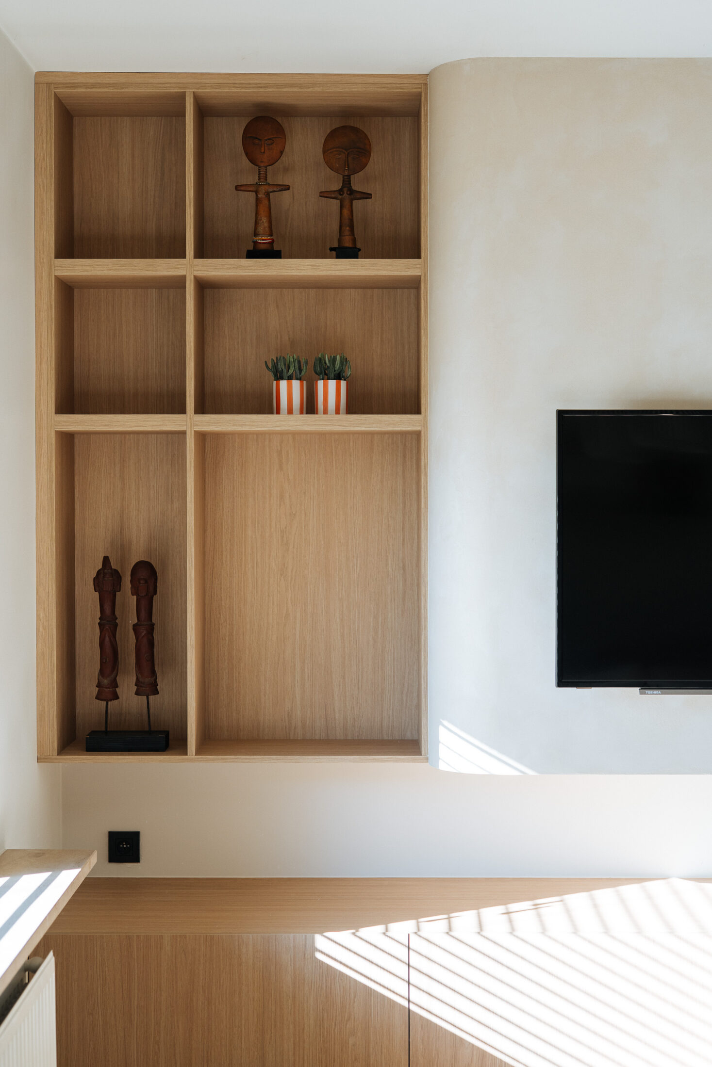Tv-meubel op maat in lichte eikenlook met afgeronde hoek en open vakkenkast naast een beige tv-wand, Kaster Keukens.