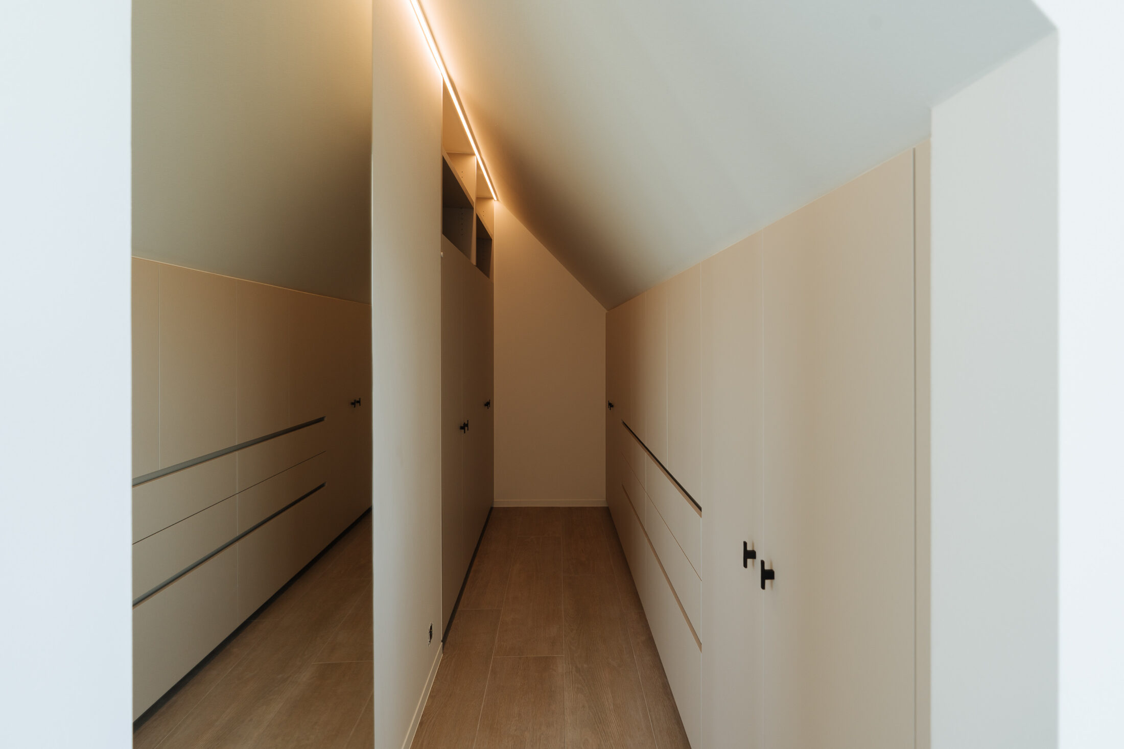 Dressing op maat onder een schuin dak met beige kasten, lades, een grote spiegel en geïntegreerde verlichting, Kaster Keukens.