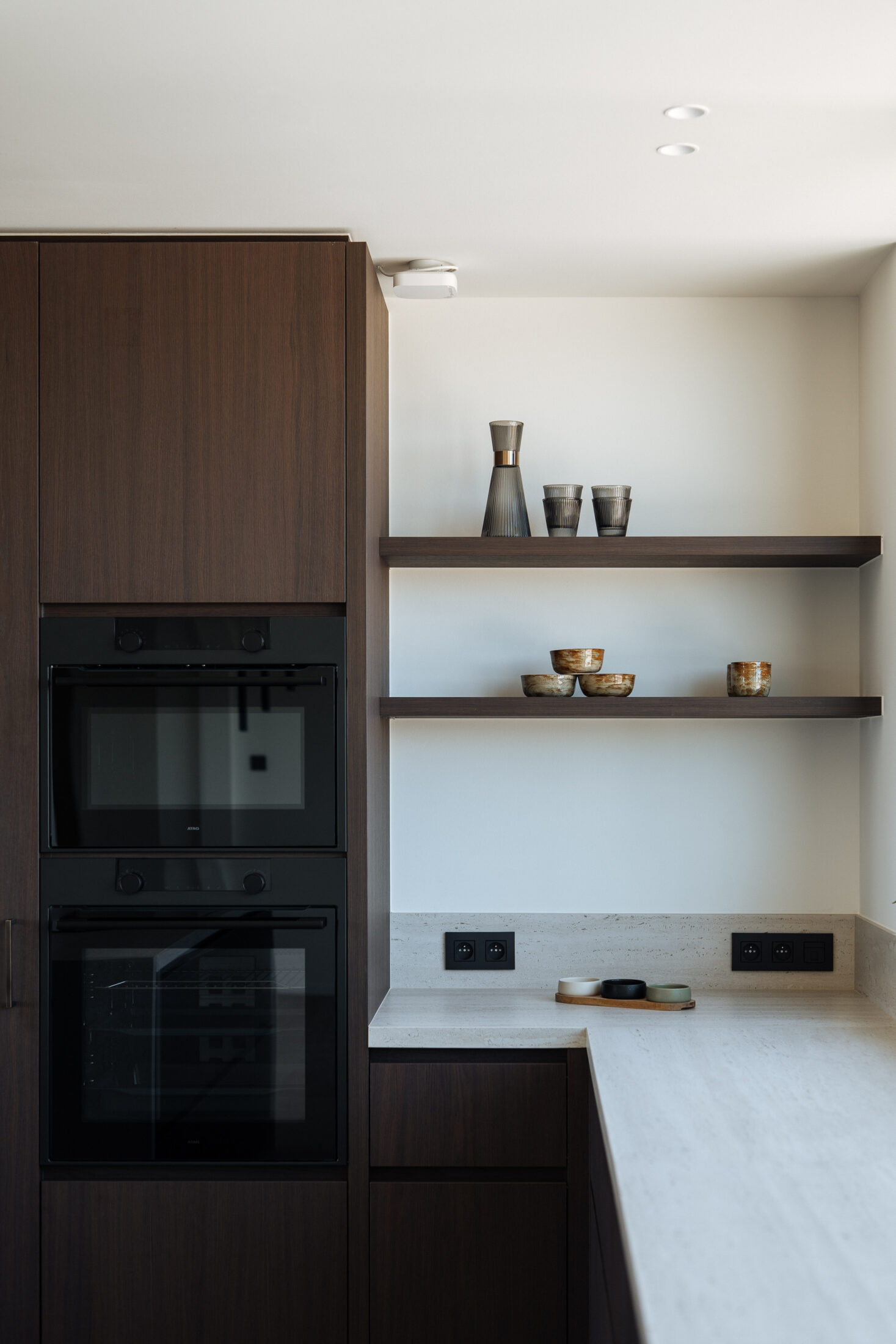 Moderne keuken met donkere houtlook fronten, licht werkblad met doorlopende zijwanden, ruim kookeiland en zwarte hanglampen, Kaster Keukens.