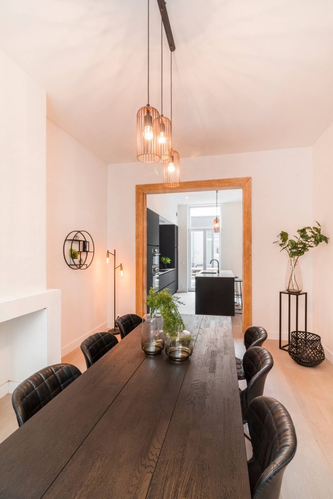 Industriële keuken gezien vanuit de eetkamer met lange houten tafel en koperen hanglampen in Gent, Kaster Keukens