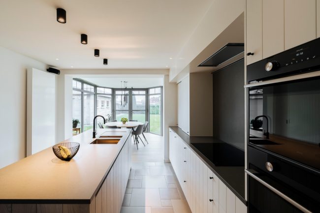 Landelijke keuken met eiken eiland, witte kasten en zwarte ovens in een lange open ruimte in Dadizele, Kaster Keukens