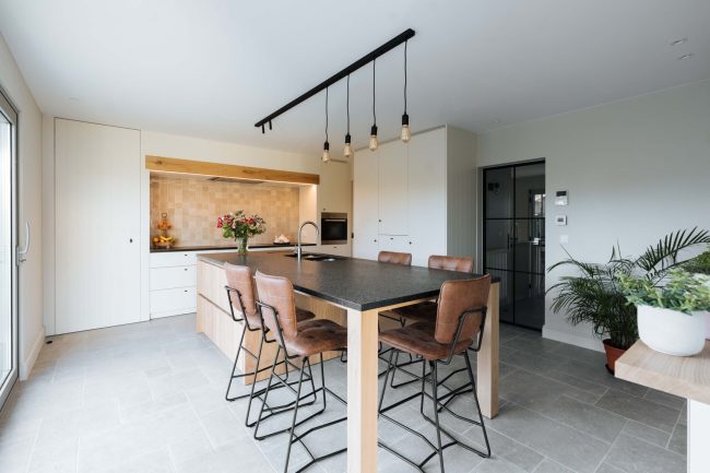 Landelijke keuken met eiken eiland, donkere tafel, bruine barkrukken en hanglampen in Ieper, Kaster Keukens