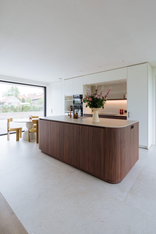 Moderne keuken met afgerond notelaar eiland in een open leefruimte met grote ramen in Hooglede, Kaster Keukens