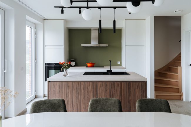 Moderne keuken met witte kasten, notelaar eiland en zwarte hanglampen naast de trap in Meulebeke, Kaster Keukens