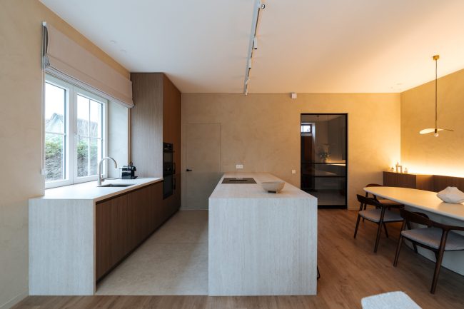Moderne keuken met licht natuurstenen eiland, notelaar kasten en open eetruimte in Oostduinkerke, Kaster Keukens
