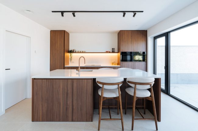 Moderne keuken met notelaarlook fronten, wit composiet werkblad, koperkleurige kraan en lichte barkrukken met houten onderstel, Kaster Keukens.
