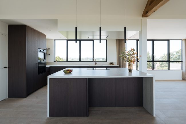 Moderne keuken met donkere houtlook fronten, licht werkblad met doorlopende zijwanden, ruim kookeiland en zwarte hanglampen, Kaster Keukens.