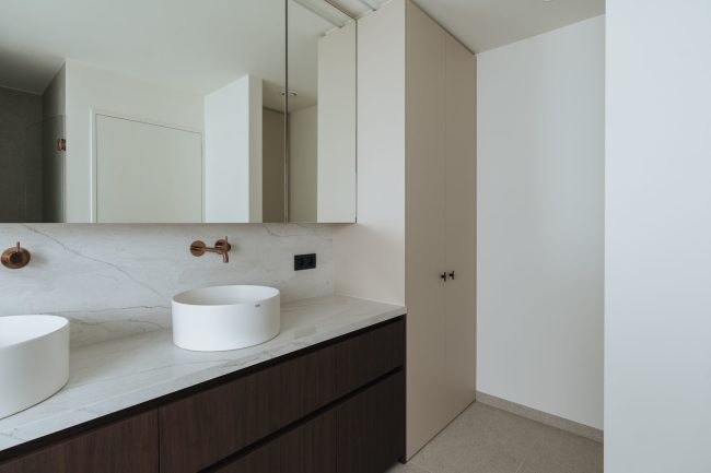 Badkamermaatwerk met donkere houtlook fronten, licht geaderd blad, witte opzetwaskommen en een hoge beige kast, Kaster Keukens.
